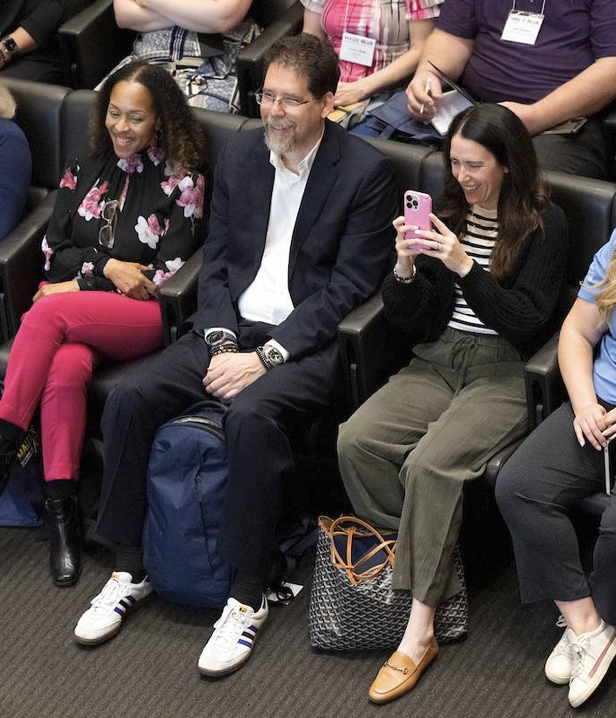 Three people sit in auditorium seats; one is holding a smartphone and taking a photo.