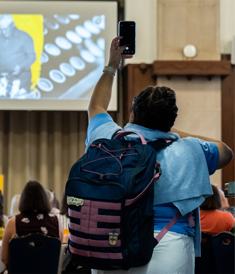An attendee holds her phone up to take a picture of the projection screen during a presentation.