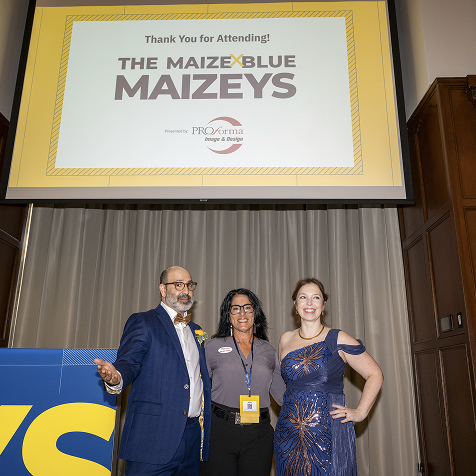 Three people standing on a stage underneath a screen that says The Maize & Blue Maizeys.